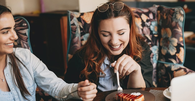 コーヒーショップで彼女の女性の友人とチーズケールを食べながら目を閉じて笑っている美しい若いプラスサイズの女性の肖像画。