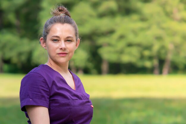 Portrait of beautiful young serious attractive professional woman in purple uniform, happy female doctor outdoors looking at camera on natural sunny green background. Copy space, place for text.