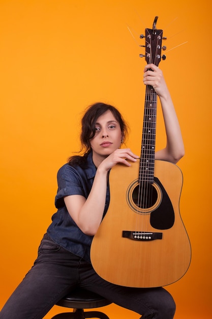 Ritratto di giovane e bella cantante che tiene la sua chitarra acustica in studio su sfondo giallo. donna abbastanza caucasica con uno strumento musicale.