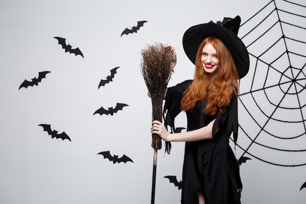 Portrait of beautiful young witch with broomstick over grey wall with bat and spider web background.