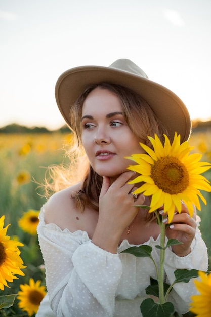 Portrait of beautiful young woman 33 years old in hat in sunflower field at sunset Happy model in white dress on summer evening in nature Warm