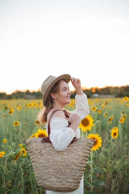 Portrait of beautiful young woman 33 years old in hat in sunflower field at sunset Happy model in white dress on summer evening in nature Warm