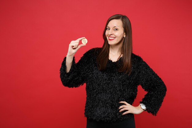 Portrait of beautiful young woman in black fur sweater holding bitcoin, future currency isolated on bright red wall background in studio. People sincere emotions lifestyle concept. Mock up copy space.