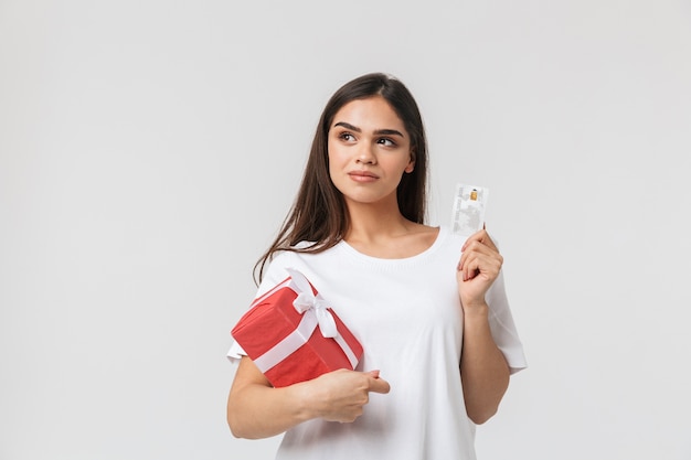 Portrait of a beautiful young woman casualy dressed standing isolated on white, holding gift box, showing credit card