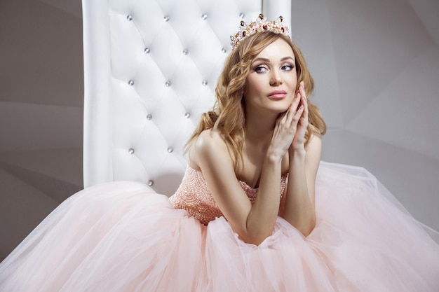 Portrait of beautiful young woman in crown and luxurious pink dress posing on stylish white armchair. sitting, touching her face and looking away. indoor studio shot.