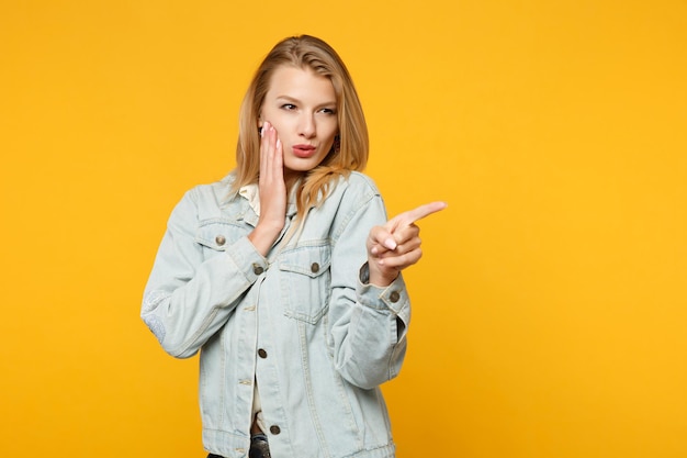 人差し指を脇に向けて、デニムのカジュアルな服を着た美しい若い女性の肖像画、スタジオで黄色のオレンジ色の背景に分離された頬に手を置きます。人々のライフスタイルのコンセプト。コピースペースをモックアップします。