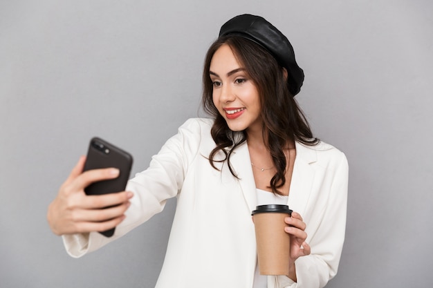 Ritratto di una giovane e bella donna vestita in giacca su sfondo grigio, tenendo la tazza di caffè, prendendo un selfie con il telefono cellulare