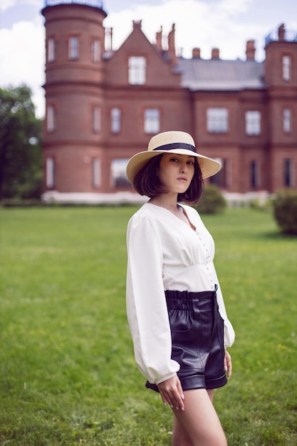 Portrait of a beautiful young woman in a hat and a white blouse and shorts goes on a green meadow in summer against the background of a large castle building