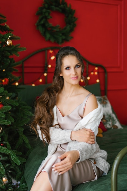 Portrait of a beautiful young woman in home clothes at the Christmas tree, she is sitting on the bed