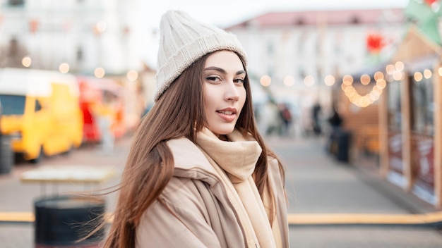 Il ritratto di una giovane e bella donna con un cappello lavorato a maglia con una giacca beige e una sciarpa cammina per la città