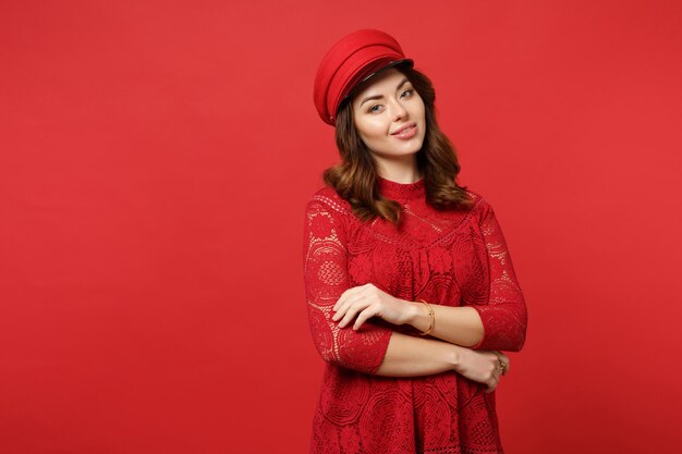 Portrait of beautiful young woman in lace dress and cap looking camera holding hands folded isolated on bright red background in studio. People sincere emotions, lifestyle concept. Mock up copy space.