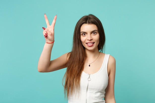 Portrait of beautiful young woman in light casual clothes showing tongue, victory sign isolated on blue turquoise background in studio. People sincere emotions, lifestyle concept. Mock up copy space.