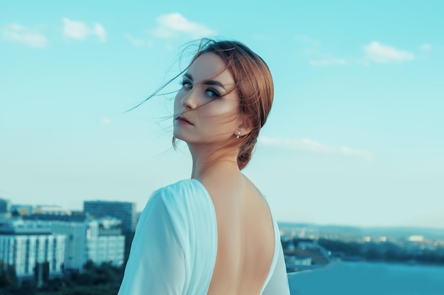 Foto ritratto di giovane e bella donna in abito bianco lungo in piedi sul tetto di un edificio con paesaggio urbano