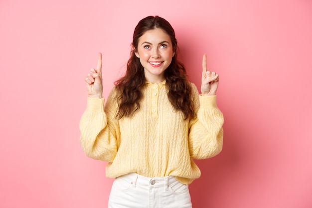 Portrait of beautiful young woman smiling, pointing and looking up at top promo offer, showing advertisement, reading promotional text, standing against pink wall.