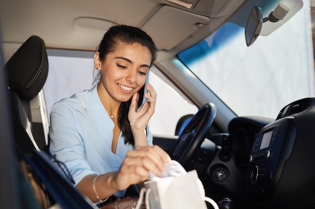 Portrait of beautiful young woman speaking by smartphone in car and smiling while looking in shopping bags, copy space