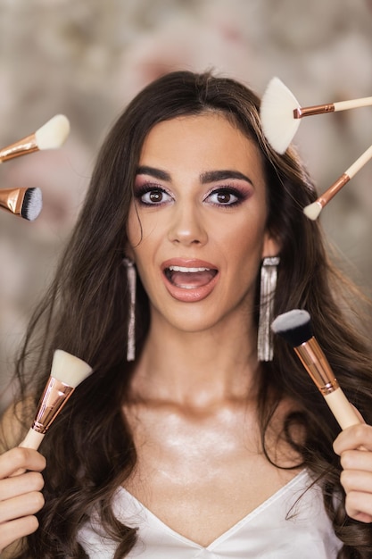 A portrait of a beautiful young woman surrounded by makeup brushes who is presenting a nice makeup on her face.