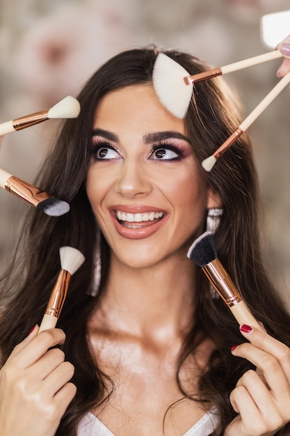 A portrait of a beautiful young woman surrounded by makeup brushes who is presenting a nice makeup on her face.