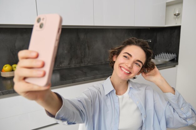 Portrait of beautiful young woman taking selfie on mobile phone at home poses for photo with