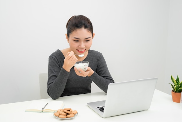 彼女のラップトップを使用して、家でヨーグルトを食べる美しい若い女性の肖像画。