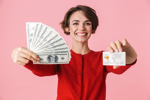 ピンクの背景の上に孤立して立っている赤い服を着て、お金の紙幣を表示し、クレジットカードを保持している美しい若い女性の肖像画