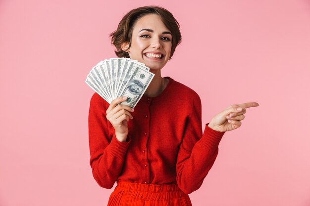 Portrait of a beautiful young woman wearing red clothes standing isolated over pink background, showing money banknotes