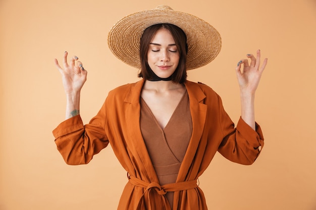 Portrait of a beautiful young woman wearing straw hat standing isolated over beige wall, meditating