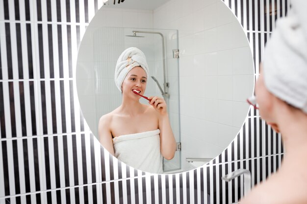 Portrait of a beautiful young woman wearing white towel brushing teeth in front of her bathroom mirror Morning routine beauty procedure