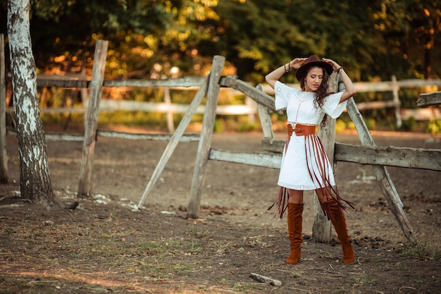 Portrait of a beautiful young woman in a white dress with over\
the knee boots and a hat with a belt and a fringe on a rustic farm\
stable