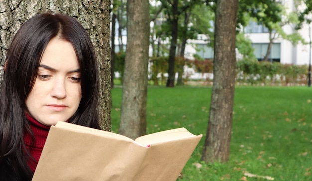 木の下に座って、楽しい晴れた日に緑の芝生の上の都市公園で彼女のお気に入りの本を読んでいる美しい若い女性の肖像画。休暇、教育、研究の概念。