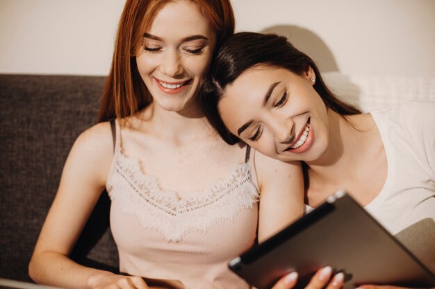 Photo portrait of a beautiful young woman with black long hair laughing while looking at her tablet screen leaning head on her female friend with red hair.