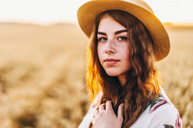 Portrait of a beautiful young woman with curly hair and
freckles face. woman in dress and hat posing in wheat field at
sunset and looking