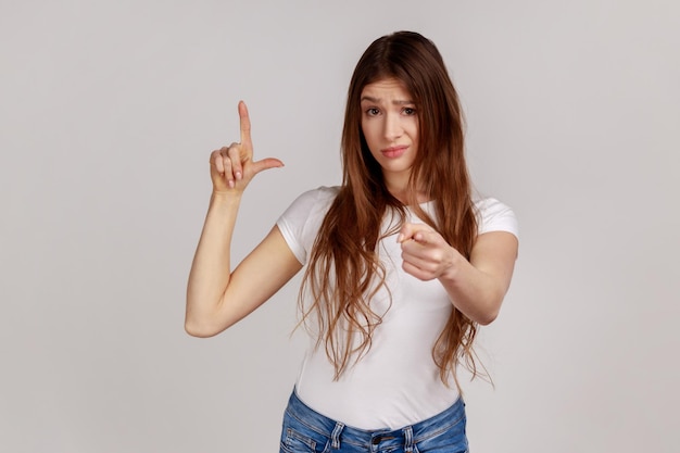 カメラを指して、白いTシャツを着て敗者のジェスチャーを示す黒髪の美しい若い女性の肖像画灰色の背景に分離された屋内スタジオショット