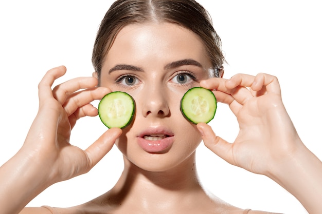 Portrait of beautiful young woman with fresh cucumber on face over white background. Concept of cosmetics, makeup, natural and eco treatment, skin care. Shiny and healthy skin, fashion, healthcare.