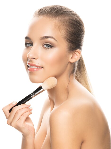 Portrait of beautiful young woman with makeup, perfect skin and bare shoulders looking at camera and holding brush. Studio shot, isolated