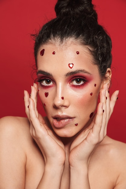 Портрет красивой молодой женщины с красным ярким макияжем, изолированным на красной стене, позирующей с сердечками на лице, кусает ее губу.