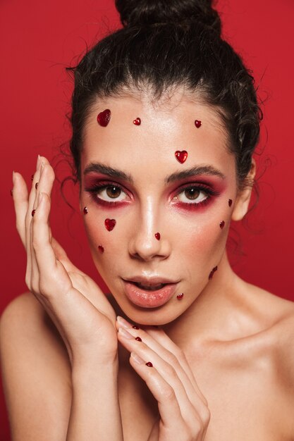 Portrait of a beautiful young woman with red bright makeup isolated on red wall posing with hearts on face.