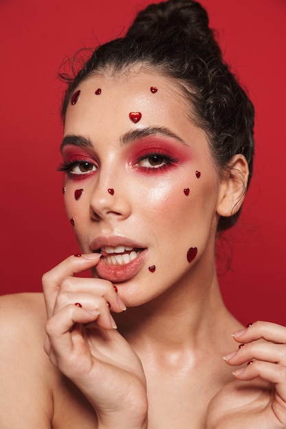 Portrait of a beautiful young woman with red bright makeup isolated on red wall posing with hearts on face.