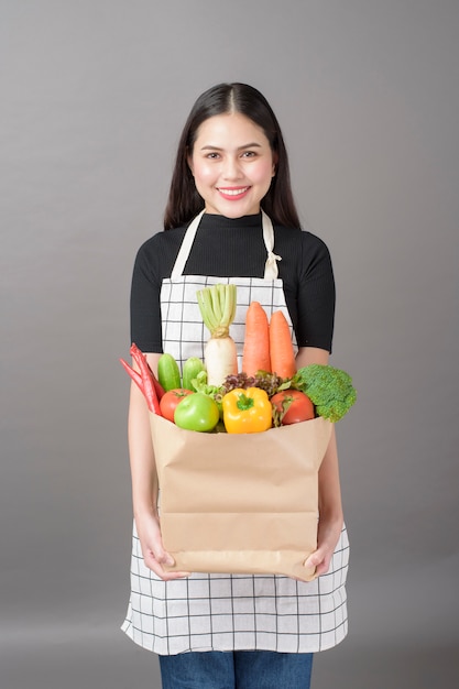 Ritratto di bella giovane donna con le verdure nella borsa di drogheria nel fondo di gray dello studio