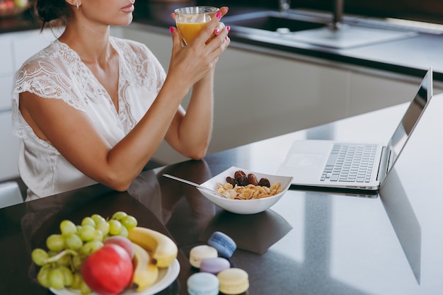 Портрет красивой молодой женщины, работающей с ноутбуком во время завтрака с хлопьями и молоком и пьющего апельсиновый сок