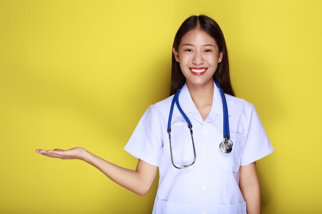 Photo portrait of a beautiful young woman in a yellow background friendly beautiful woman wearing a doctors uniform and pointing while wearing a stethoscope