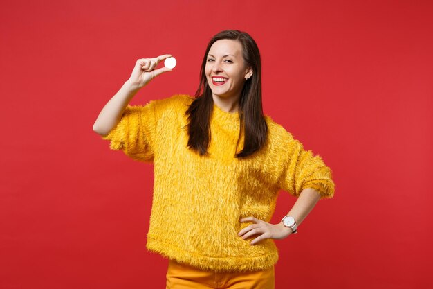 Ritratto di bella giovane donna in maglione di pelliccia gialla che tiene bitcoin valuta futura isolata su sfondo rosso brillante della parete in studio. concetto di stile di vita di emozioni sincere della gente. mock up copia spazio.