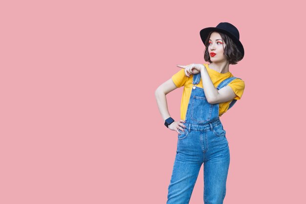 Portrait of beautiful young woman in yellow t-shirt, blue denim overalls with makeup and black hat standing with hand on waist and pointing at copypace. indoor studio shot isolated on pink background.