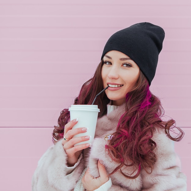 Portrait of beauty fashion smiling woman with coffee showing tongue in mink coat and black hat . Copy-space. Colored hairs