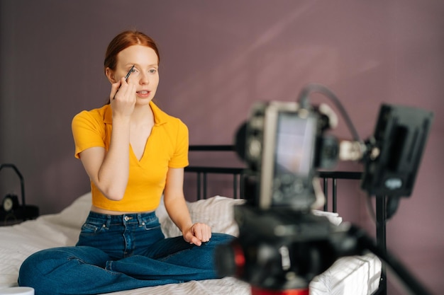 Portrait of beauty female blogger applying shadows to eyebrows with makeup brush recording daily makeup routine tutorial to professional camera at home studio