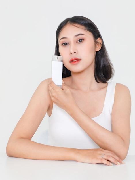 Portrait beauty studio shot millennial Asian black hair sexy female model in tank top singlet look at camera holding showing serum facial moisturizer skincare package near face on white background.