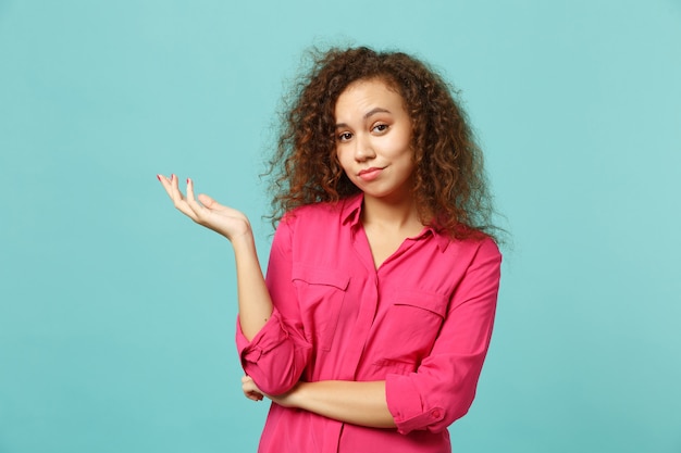 Portrait of bewildered african girl in casual clothes looking camera, spreading hands isolated on blue turquoise background in studio. People sincere emotions, lifestyle concept. Mock up copy space.