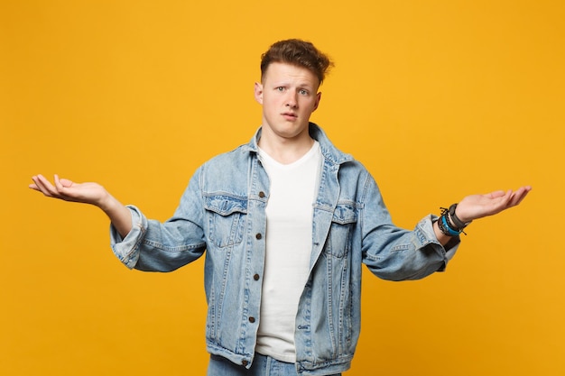 Portrait of bewildered upset young man in denim casual clothes pointing spreading hands aside isolated on yellow orange wall background. People sincere emotions, lifestyle concept. Mock up copy space.