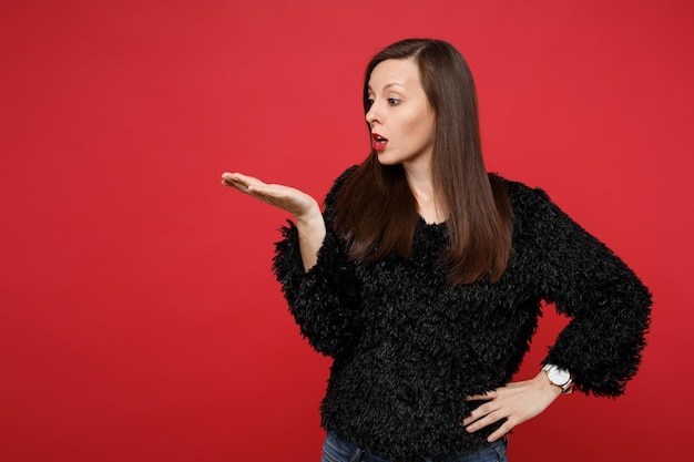 Portrait of bewildered young woman in black fur sweater looking, pointing hand aside isolated on bright red wall background in studio. People sincere emotions, lifestyle concept. Mock up copy space.