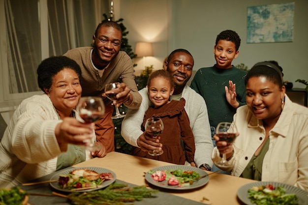 Ritratto di grande famiglia afroamericana che alza i bicchieri alla telecamera mentre si gode la cena di natale insieme...