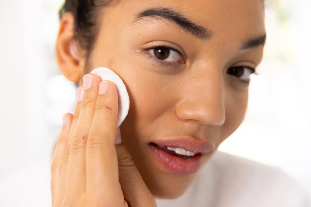 Photo portrait of biracial woman in bathrobe cleansing her face in sunny bathroom